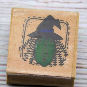 May include: A wooden rubber stamp with a witch's face and hat design. The witch has a green face and a black hat with a blue band. The stamp is surrounded by a decorative border.