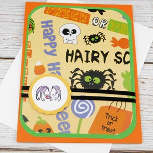 May include: Halloween greeting card with an orange border and green trim. The card features a yellow background with Halloween-themed graphics, including spiders, skulls, and the words "Trick or Treat" and "Happy Halloween". A round sticker with a purple design is in the center.