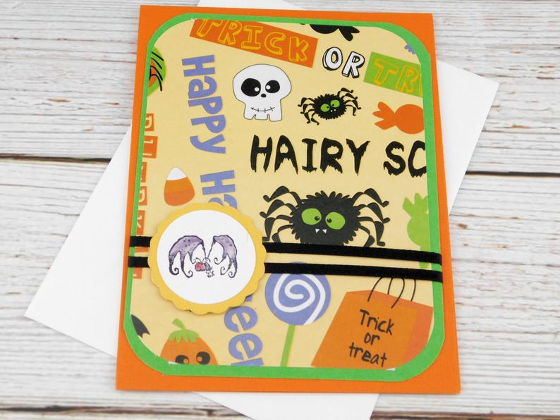 May include: Halloween greeting card with an orange border and green trim. The card features a yellow background with Halloween-themed graphics, including spiders, skulls, and the words "Trick or Treat" and "Happy Halloween". A round sticker with a purple design is in the center.