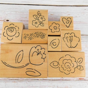 May include: A collection of wooden stamps with various floral designs. The stamps feature outlines of flowers, leaves, and swirls in black ink. The blocks are arranged on a light wooden surface.