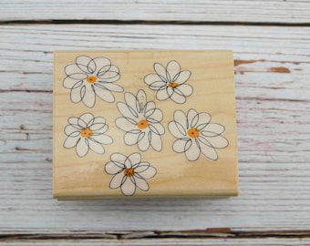 May include: A wooden stamp featuring a daisy design. The stamp has a light beige background with six daisy illustrations. Each daisy has white petals, black outlines, and orange centers. The stamp is rectangular.