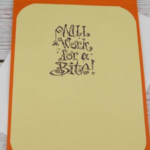 May include: A greeting card with an orange border and a light yellow interior. The card features the phrase "Will Work for a Bite!" in dark purple, stylized lettering. A white envelope is tucked inside.