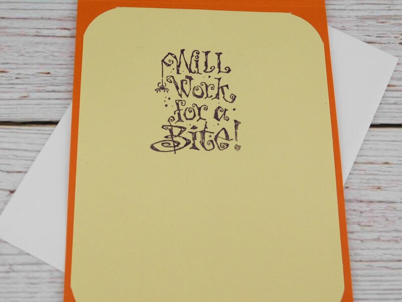 May include: A greeting card with an orange border and a light yellow interior. The card features the phrase "Will Work for a Bite!" in dark purple, stylized lettering. A white envelope is tucked inside.