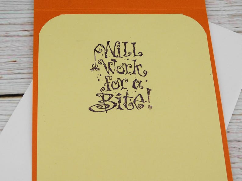 May include: A greeting card with an orange border and a light yellow interior. The text "Will Work for a Bite!" is written in a whimsical, dark brown font, with a spider graphic.