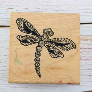 May include: A wooden stamp with a black and white detailed dragonfly design. The dragonfly has intricate patterns on its wings and body.