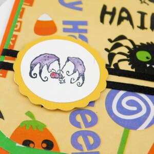 May include: A Halloween-themed greeting card with a light yellow background. The card features a circular image of two purple bats, a black spider, and the word "HAIRY". Other elements include a candy corn, a lollipop, and a pumpkin.