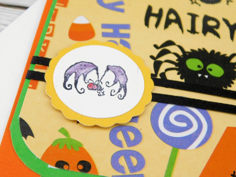 May include: A Halloween-themed greeting card with a light yellow background. The card features a circular image of two purple bats, a black spider, and the word "HAIRY". Other elements include a candy corn, a lollipop, and a pumpkin.