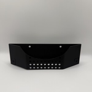 May include: A black, wall-mounted storage basket with a rectangular shape and a series of small, evenly spaced holes along the bottom. The basket has a curved front and two mounting holes.