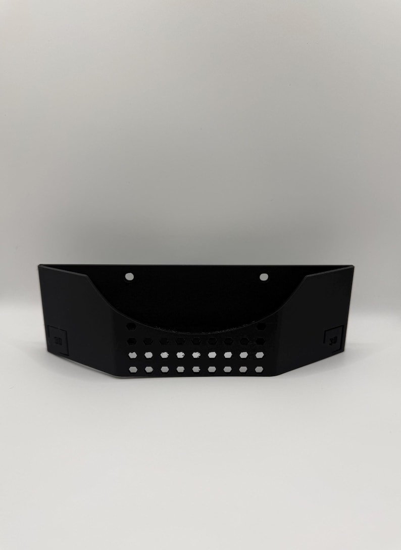 May include: A black, wall-mounted storage basket with a rectangular shape and a series of small, evenly spaced holes along the bottom. The basket has a curved front and two mounting holes.