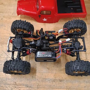 Pode incluir: Um camião vermelho desmontado com controlo remoto, com chassis preto e pneus grandes e com saliências. A carroçaria vermelha do camião está separada, com um autocolante de estrela branca. Os componentes internos do camião, incluindo a bateria, são visíveis.