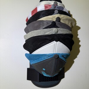 May include: A stack of baseball caps in various colors and designs, including black, white, gray, and blue. The caps are displayed on a black wall-mounted rack, showcasing their different styles and patterns.
