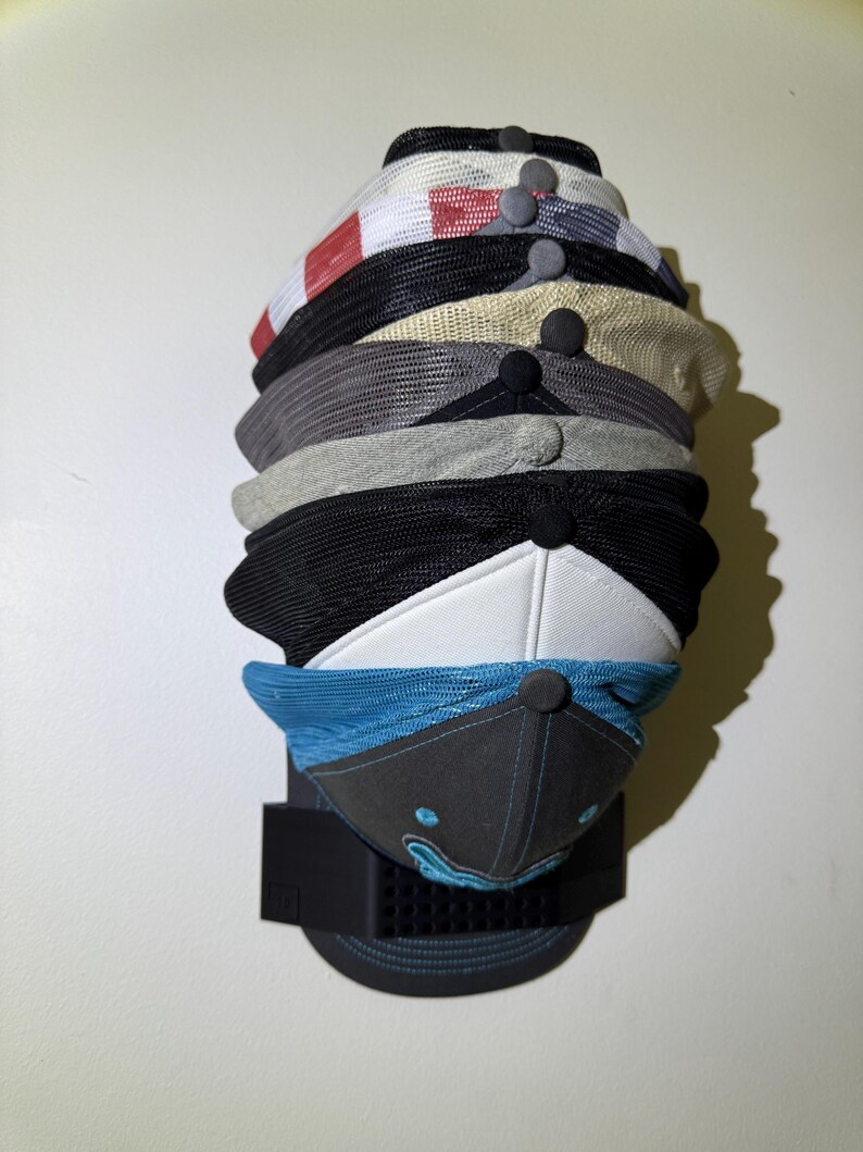 May include: A stack of baseball caps in various colors and designs, including black, white, gray, and blue. The caps are displayed on a black wall-mounted rack, showcasing their different styles and patterns.