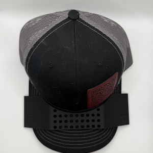 May include: Black and gray trucker hat with a mesh back and a black front panel. The hat features a red leather patch with a QR code. The hat is displayed on a black hat holder.