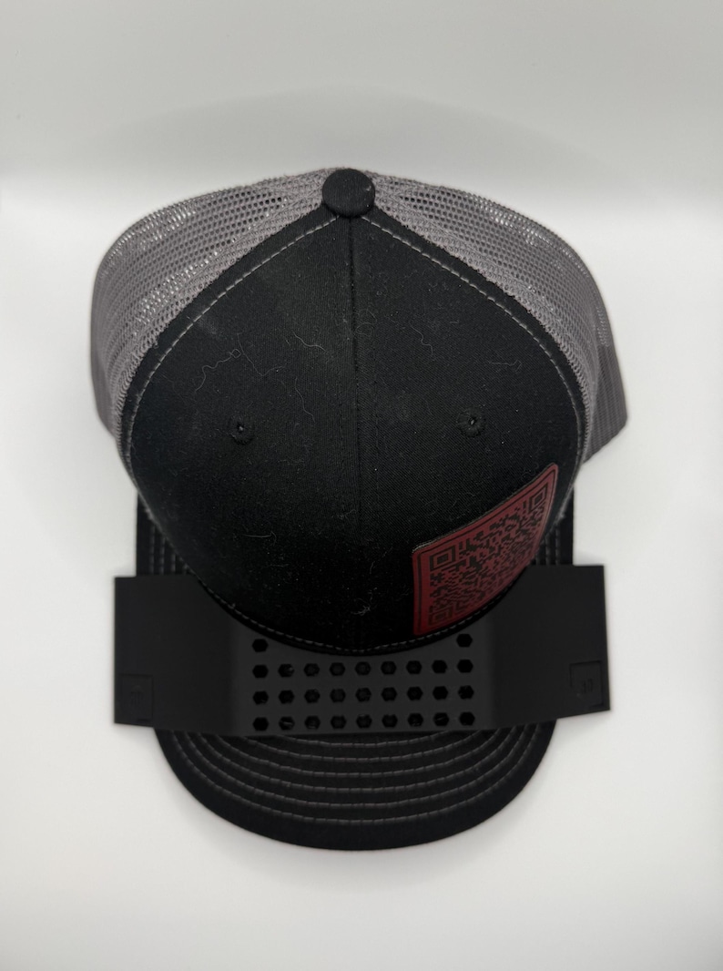 May include: Black and gray trucker hat with a mesh back and a black front panel. The hat features a red leather patch with a QR code. The hat is displayed on a black hat holder.