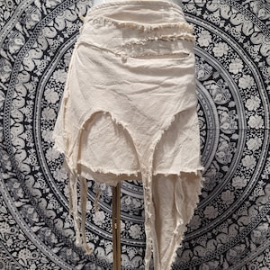 May include: A cream-colored skirt featuring a layered design with raw, frayed edges and uneven, jagged hemlines. The garment has a wrap-style construction with multiple fabric panels that hang at varying lengths, creating a distressed appearance.