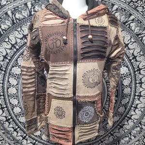 May include: A zip-up hooded jacket featuring a patchwork design in shades of brown, tan, and black. The garment includes distressed horizontal slits, visible stitching, and printed graphics including an Om symbol and a sun face design.