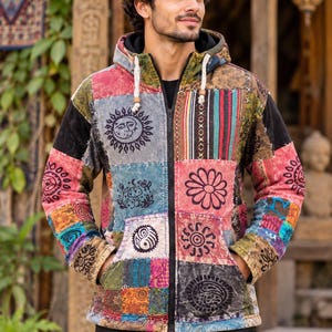 Op de afbeelding: Een jack met rits en capuchon met een patchworkontwerp van diverse kleurrijke stoffen vierkanten. De patronen bevatten zwarte prints van zonnen, bloemen, paddenstoelen en paisley-motieven. Het jack heeft een zwarte ritssluiting aan de voorzijde.