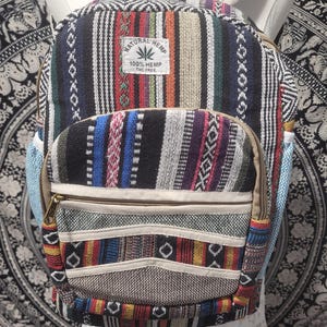 May include: A backpack featuring a multi-coloured woven pattern with stripes and geometric shapes. It includes a front zippered pocket and a white rectangular label that reads Natural Hemp 100% Hemp THC Free. The bag has beige straps and zippers.