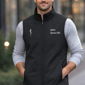 May include: Black vest with a zipper closure, two side pockets, and a stand-up collar. The vest has a white medical symbol on the left chest and the embroidered text "John James DO" on the right chest. The person is wearing a gray long-sleeve shirt underneath.
