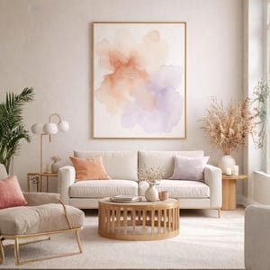 May include: A living room scene featuring a large abstract watercolor painting in a gold frame. A cream-colored sofa with peach and lavender pillows sits below the artwork. A round wooden coffee table and a gold-framed armchair complete the decor.