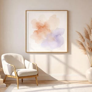 May include: A framed watercolor abstract art print with soft orange, peach, and purple hues. The artwork is displayed in a gold frame, complementing the neutral tones of the room. A white armchair with gold accents and a vase of dried pampas grass complete the decor.