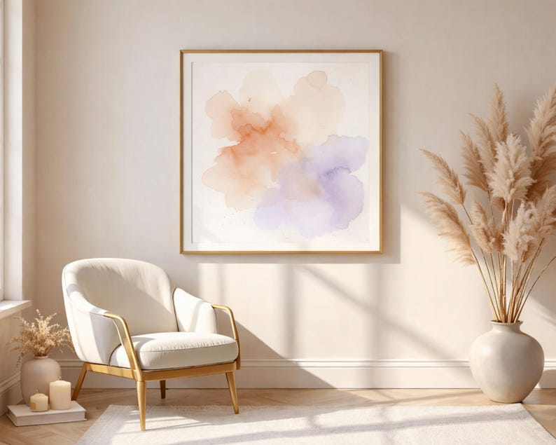 May include: A framed watercolor abstract art print with soft orange, peach, and purple hues. The artwork is displayed in a gold frame, complementing the neutral tones of the room. A white armchair with gold accents and a vase of dried pampas grass complete the decor.