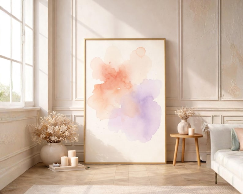 May include: A large framed watercolor art print featuring a soft, abstract design in shades of peach, coral, and lavender. The artwork is displayed in a bright, airy room with a neutral color palette, complemented by a wooden side table and a white sofa.