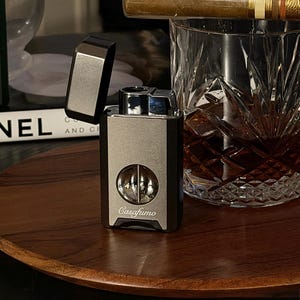 May include: A metallic gray lighter with the text "Casafumo" is open, revealing its interior. A cigar rests on a crystal rocks glass filled with amber liquid. The items are on a wooden tray.