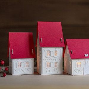 Christmas village from side angle showing white detail. White houses, red roofs. Smallest house has a small, stone-textured chimney made of paper. Holiday greens along side them.