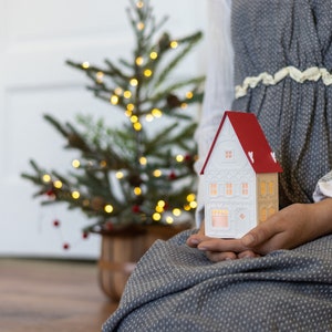 Chrstmas village house being held in the hands of a young woman. House is white, with white details, and a red roof. Candle twinkles inside it. Small Christmas tree in background.