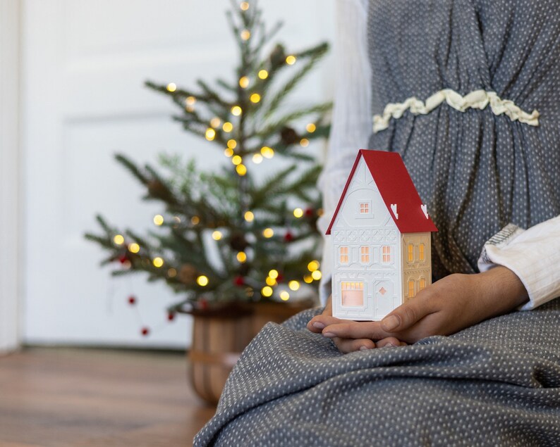 Chrstmas village house being held in the hands of a young woman. House is white, with white details, and a red roof. Candle twinkles inside it. Small Christmas tree in background.