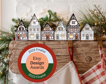 May include: A row of six miniature wooden houses with black, white, and brown details, arranged on a wooden beam. The houses have varying architectural designs. A round sticker with "Etsy Design Awards 2023 Finalist" is in the foreground.