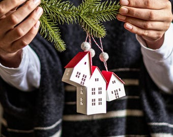 Puede incluir: Tres casas de papel blanco con techos rojos cuelgan de una rama de pino. Las casas están decoradas con pequeñas ventanas y están unidas con un cordel blanco.