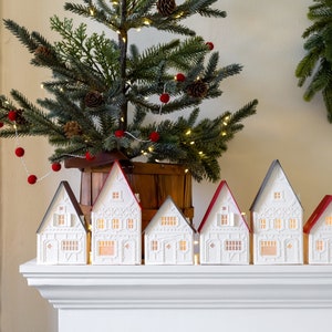 White Christmas village with red and gray roofs on white mantel. Small Christmas tree behind them. Small wreath hanging on the wall behind them.