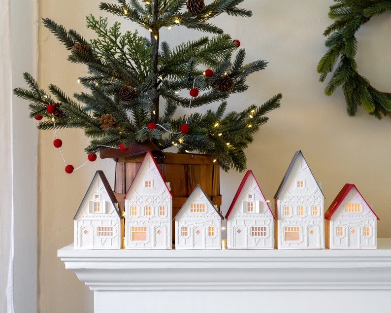 White Christmas village with red and gray roofs on white mantel. Small Christmas tree behind them. Small wreath hanging on the wall behind them.