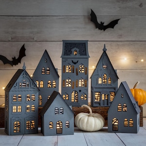 May include: A collection of gray, illuminated paper houses with intricate window designs, arranged on a wooden surface. The scene is decorated with paper bats and pumpkins, creating a Halloween theme. Warm light shines from within the houses.