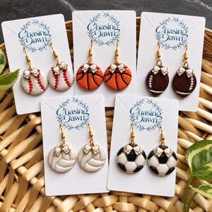 May include: Five pairs of dangle earrings on white display cards labeled Chasing Dawn. Each pair features a sports ball design including baseball, basketball, football, volleyball, and soccer ball motifs, all accented with small white bows and gold hooks.