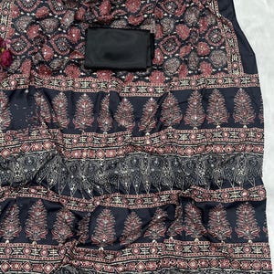 May include: A black textile featuring detailed patterns in red, cream, and gold. The design incorporates floral and geometric motifs, with a solid black rectangular piece on top. The fabric is draped, highlighting the intricate print.