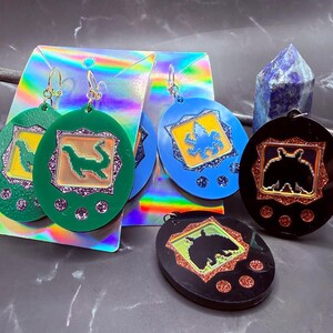 May include: Earrings in green, blue, and black, shaped like retro handheld game consoles. Each earring features a different design inside, with glitter accents and gold-colored hooks. The earrings are displayed on holographic cards.