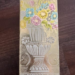 May include: A rectangular, gold-coloured box with a floral design. The embossed design features a white vase filled with colourful flowers in shades of pink, blue, and yellow. The box has a textured surface and a brown top.