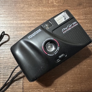 May include: Black Keystone Easy Shot 500X camera with electronic auto wind. The camera has a 38mm lens and a built-in flash. The camera has a black strap attached. The camera is on a brown wooden surface.