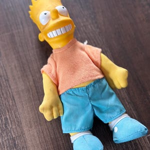 May include: A Bart Simpson plush doll with a yellow head and arms, an orange shirt, and blue shorts and shoes. The doll has a wide grin and white teeth. The doll is lying on a brown wooden surface.