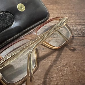 May include: A pair of eyeglasses with a light brown frame and clear lenses, resting next to a black leather case. The glasses have a rectangular shape and a gold-colored hinge. The case has a gold-colored emblem that says "YES".