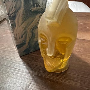 May include: A vintage amber-colored glass perfume bottle in the shape of a stylized face with a mohawk-like design. The bottle sits on a wooden surface, with a box featuring a bas-relief of a horse and rider in the background.