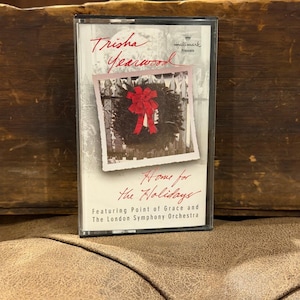 Könnte beinhalten: Eine Kassettenhülle mit dem Album Home for the Holidays von Trisha Yearwood, Hallmark Presents. Das Cover zeigt ein Foto eines grünen Kranzes mit roter Schleife an einem weißen Zaun sowie Text, der Point of Grace und das London Symphony Orchestra nennt.