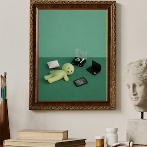 May include: A framed artwork featuring a green figure, pillow, miniature laptop, record player, and tablet on a green background. The frame is ornate with a brown finish. A bust and books are on a wooden surface.