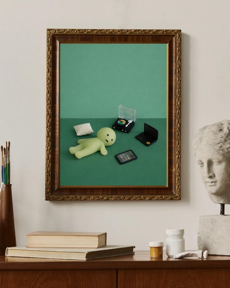 May include: A framed artwork featuring a green figure, pillow, miniature laptop, record player, and tablet on a green background. The frame is ornate with a brown finish. A bust and books are on a wooden surface.