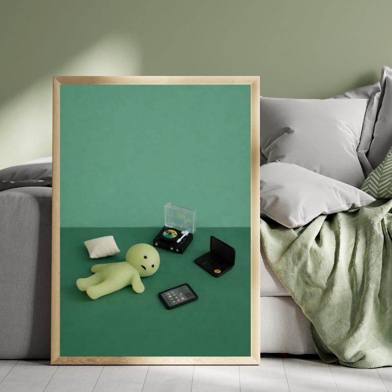 May include: A framed print featuring a green figure lying down with a small pillow, a miniature record player, a laptop, and a tablet on a green surface. The background is a solid green color, and the frame is a light wood color.
