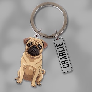 Pode incluir: Um porta-chaves de metal prateado com uma ilustração de um cão pug bege e uma etiqueta retangular branca com o texto preto CHARLIE. O pug é representado numa posição sentada com um focinho preto e olhos escuros, num anel circular.