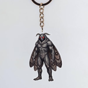 May include: A keychain featuring a clear acrylic charm of a muscular, humanoid moth creature with dark gray skin, large wings, and glowing red eyes. The charm is attached to a circular metal ring by a short silver-toned metal chain link.